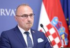 Muški govornik u plavom odijelu i naočalama uz hrvatsku zastavu tijekom službenog obraćanja