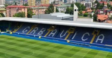 Pogled na praznu Grbavicu sa zelenim terenom i plavim banderolama FK Sarajeva, grad u pozadini