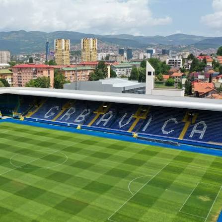 Pogled na praznu Grbavicu sa zelenim terenom i plavim banderolama FK Sarajeva, grad u pozadini