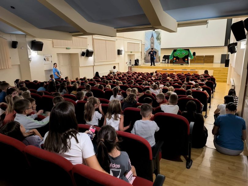Većina djece u dvorani sjedí i gleda predstavu na pozornici