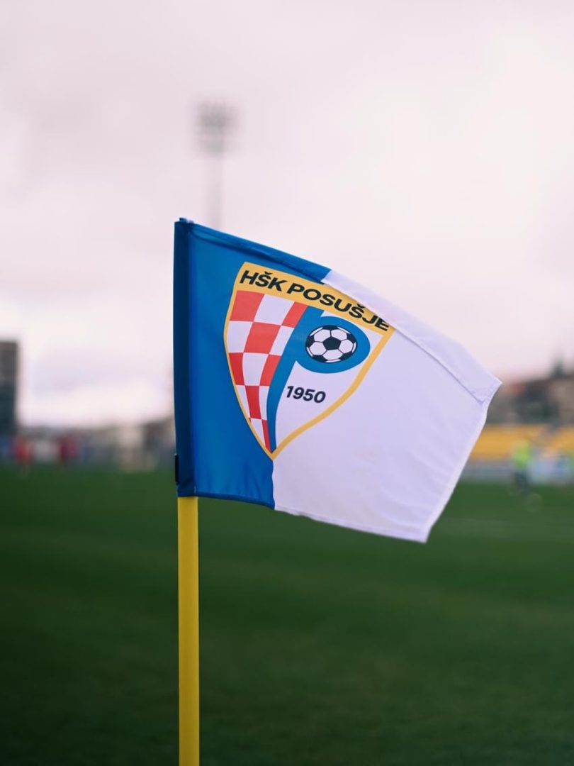 Plava i bijela zastava HŠK Posušje sa grbom na žutoj zastavici na nogometnom travnjaku