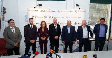 Grupa službenih osoba na konferenciji za tisak s mikrofonima ispred reklamnog panela
