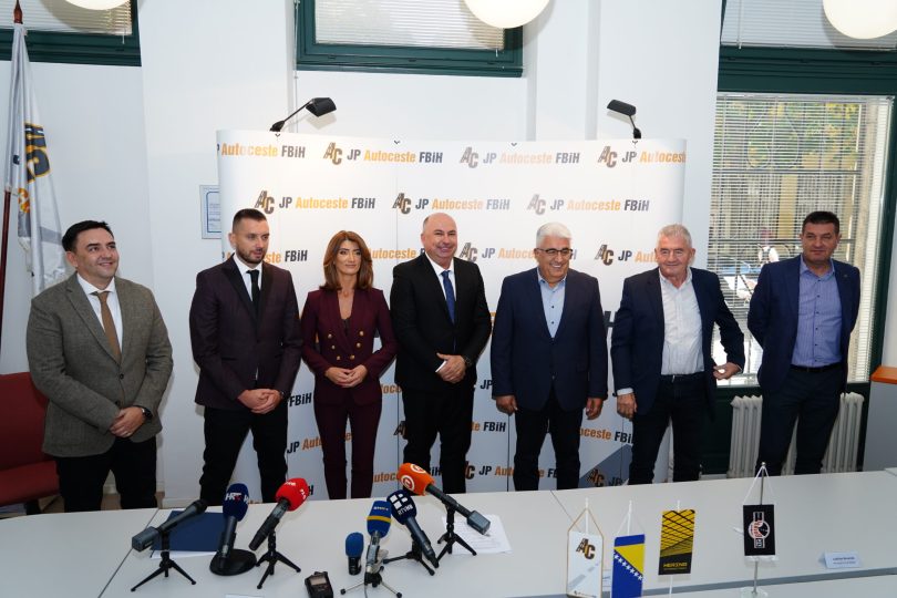 Grupa službenih osoba na konferenciji za tisak s mikrofonima ispred reklamnog panela