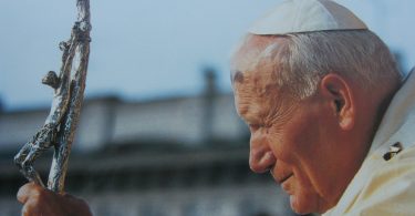 Stariji svećenik u bijeloj sutani drži drveni križ, pogleden u profilu
