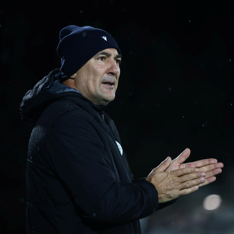 Trener u crnoj jakni i plavoj kapa govori sa gestama tijekom utakmice, noću na stadionu