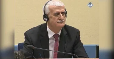 Muškarac u odijelu i kravati sjediti za stolom s mikrofonom na sudskoj sjednici
