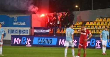 Fudbalski derbi s navijačima koji drže crvene bakljade na stadionu.