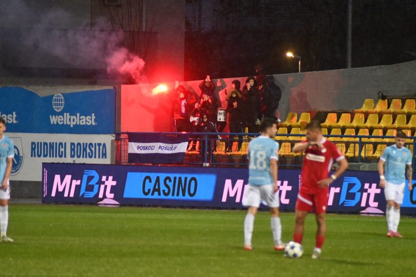 Fudbalski derbi s navijačima koji drže crvene bakljade na stadionu.