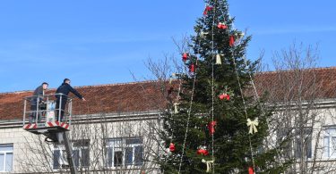 Osvijetljena Božićna jelka s crvenim украјnjицама na gradskom trgu s kamenim zgradom u pozadini