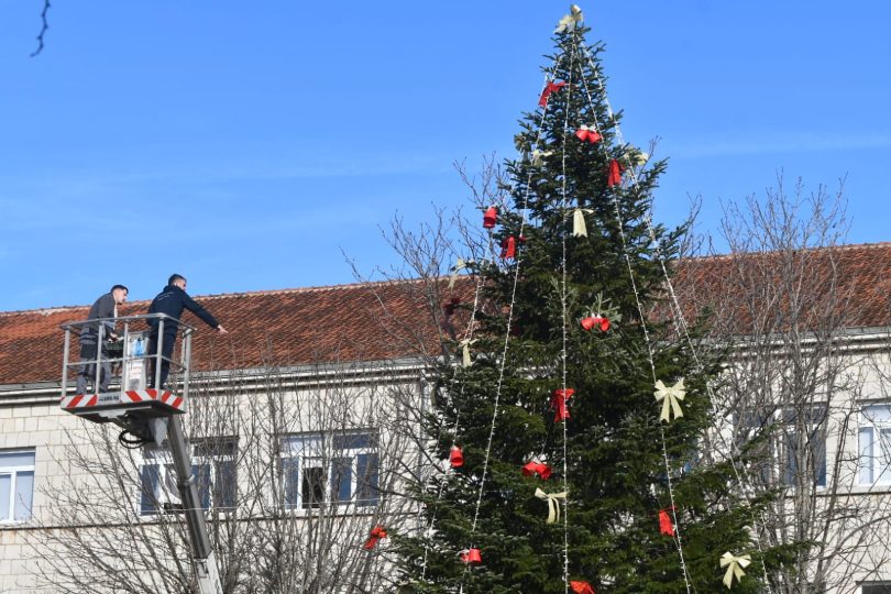 Osvijetljena Božićna jelka s crvenim украјnjицама na gradskom trgu s kamenim zgradom u pozadini