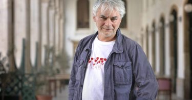 Stariji muškarac sa sivom kosom u plavoj košulji stoji u arhitektonskom prostoru