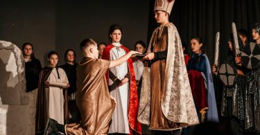 Scena iz predstave Krunidba s glumcima u historijskim kostimima, kralj sjedi na prijestolju okružen svojom povorkom
