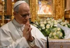 Stariji čovjek u bijeloj odjeći moli se u crkvi, s cvijećem i crkvenim ukrasima u pozadini