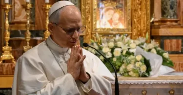 Stariji čovjek u bijeloj odjeći moli se u crkvi, s cvijećem i crkvenim ukrasima u pozadini