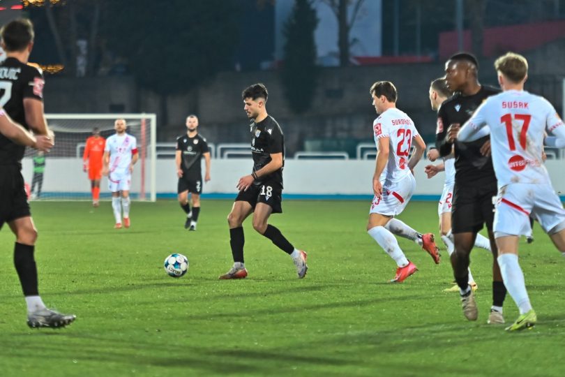 Fudbalska utakmica na terenu; igrači u crnim dresovima (Posušje) i bijelim dresovima (Sloga) u borbi za loptu