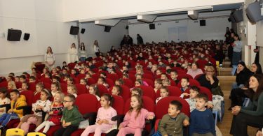 Група малишана у црвеним костимима седи у позоришту током представе.