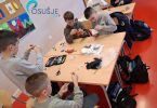Grupa učenika sjednih za stol šiva na dječjoj radionici uz pomoć odraslih instruktora