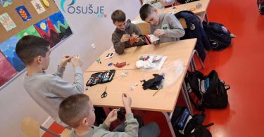 Grupa učenika sjednih za stol šiva na dječjoj radionici uz pomoć odraslih instruktora