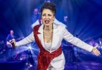 Žena u bijelom saku s crvenim detaljima na pozornici s orkestrom u pozadini