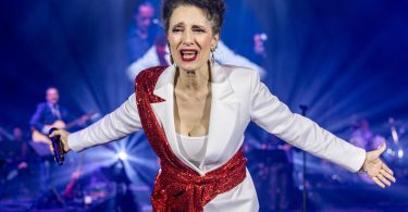 Žena u bijelom saku s crvenim detaljima na pozornici s orkestrom u pozadini