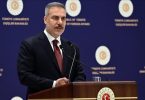 Muški govornik u tamnom odijelu i crvenoj kravati govori za mikrofonom ispred diplomskog pozadine