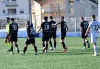 Fudbalska utakmica na terenu - igrači u crnim dresovima skupljeni oko lopte