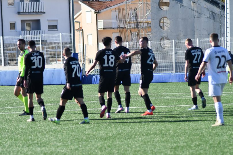Fudbalska utakmica na terenu - igrači u crnim dresovima skupljeni oko lopte