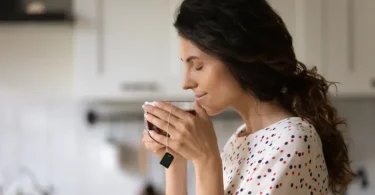 Žena s dugom crnom kosom pije toplu šalicu čaja iz bele šolje u kuhnji
