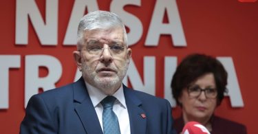 Muž u plavom odijelu i naočalama napress konferenciji uz crvenu pozadinu