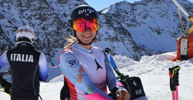 Skijašica u skijaški opremi s italijanskom zastavom na leđima, s gorama u pozadini