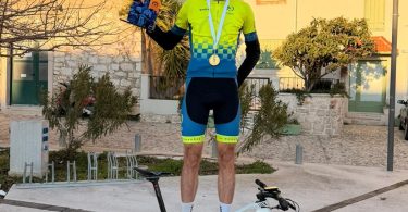 Mladić na biciklu u zeleno-plavoj majici drži trofej ispred žute kuće