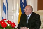 Stariji čovjek u crnom odijelu i naočalama sjedeći pred zastavama