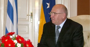 Stariji čovjek u crnom odijelu i naočalama sjedeći pred zastavama