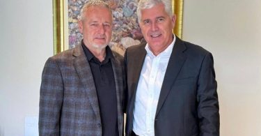 Dva muškarca poziraju za fotografiju, jedan u plavoj kockastoj jakni, drugi u crnom odijelu