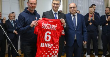 Službenik u tamnoj odjeći drži crveni rukometni dres broj 6 pored muškarca u sivom odijelu tijekom službene ceremonije