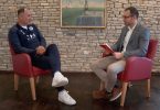 Muškarac u tamnoj majici i muškarac u sivom sakovu razgovaraju u studiju tijekom intervjua