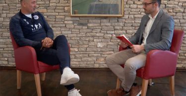 Muškarac u tamnoj majici i muškarac u sivom sakovu razgovaraju u studiju tijekom intervjua