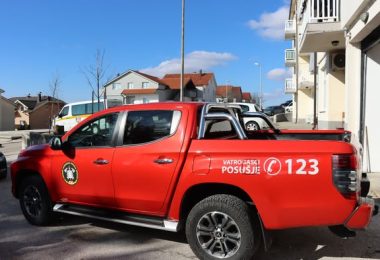 Crveno terensko vatrogasno vozilo s natpisom Vatrogasci 123 parkirano ispred zgrade