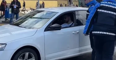 Policajac u plavoj uniformi stoji pored bijelog automobila tokom očevida na ulici