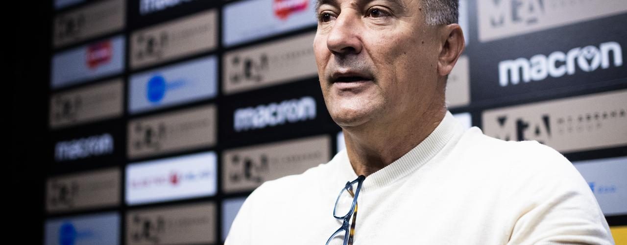Trener na press konferenciji govori u mikrofon, pozadina s logotipima sponzora