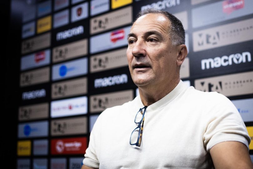 Trener na press konferenciji govori u mikrofon, pozadina s logotipima sponzora