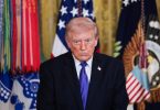 Muški političar u crnom odijelu i plavoj kravati ispred američke zastave