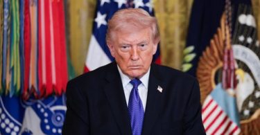 Muški političar u crnom odijelu i plavoj kravati ispred američke zastave