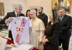 Crkveni dostojanstvenik u bijeloj odjeći gleda na crveno-bijeli sportski dres koji mu pokazuje muškarac u tamnom odijelu