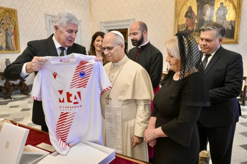 Crkveni dostojanstvenik u bijeloj odjeći gleda na crveno-bijeli sportski dres koji mu pokazuje muškarac u tamnom odijelu