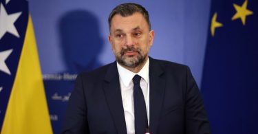 Muškarac u tamnom odijelu i kravati govori na konferenciji s pozadinom zastava BiH i EU