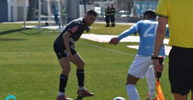 Fudbalski igrač u crvenoj majici izvršava duel s igračem u plavoj majici, sudija s žutom karticom u pozadini