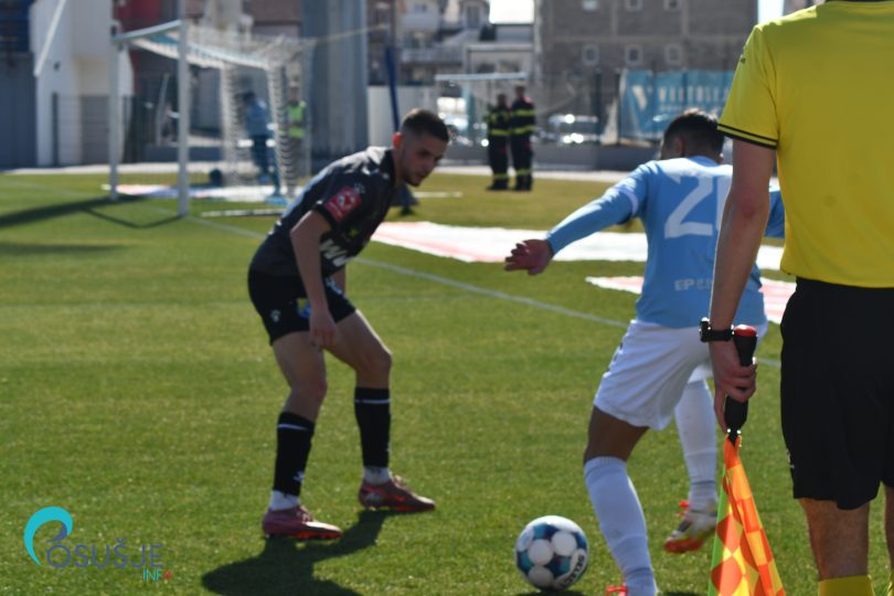 Fudbalski igrač u crvenoj majici izvršava duel s igračem u plavoj majici, sudija s žutom karticom u pozadini