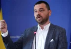 Muškarac u plavom odijelu govori na konferenciji, iza njega zastave