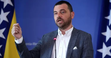 Muškarac u plavom odijelu govori na konferenciji, iza njega zastave